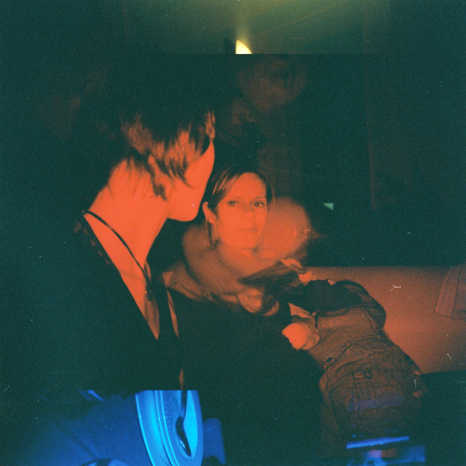 Two people sit in a dimly lit space, one facing the camera while the other looks away.