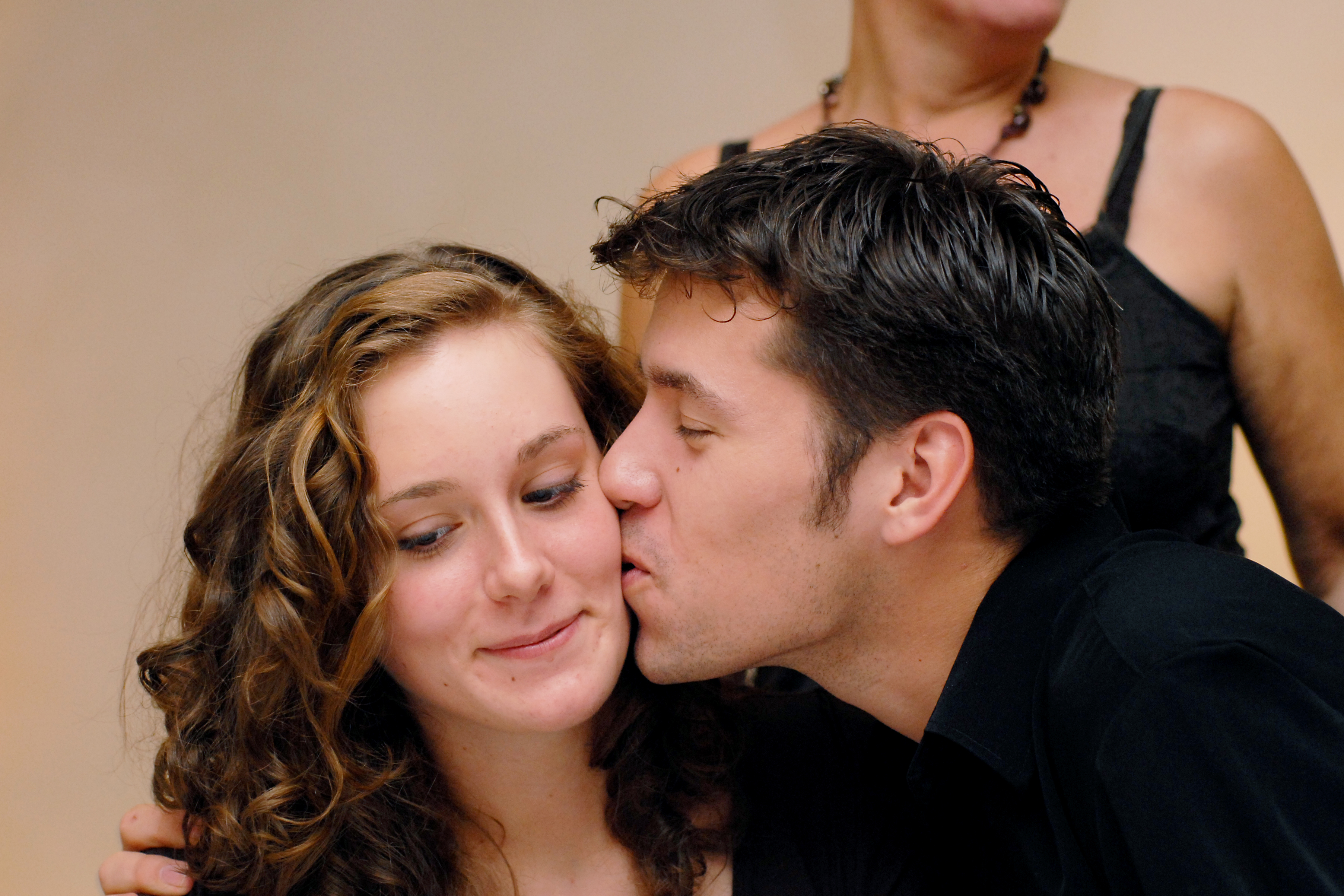 A man in a black shirt kisses a smiling woman on the cheek while another person stands in the background.