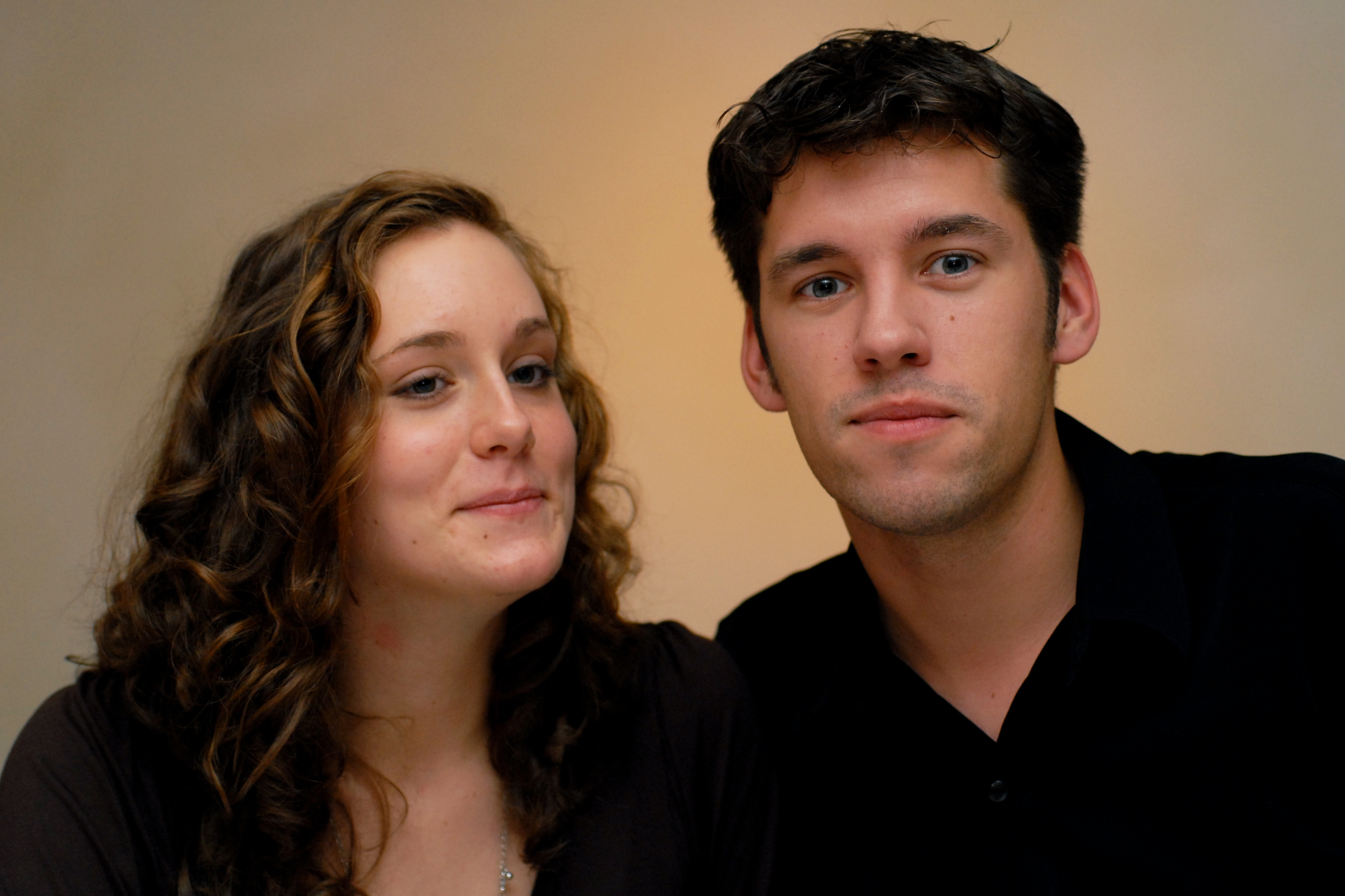 A woman with curly hair smiles slightly while a man in a black shirt looks directly at the camera.