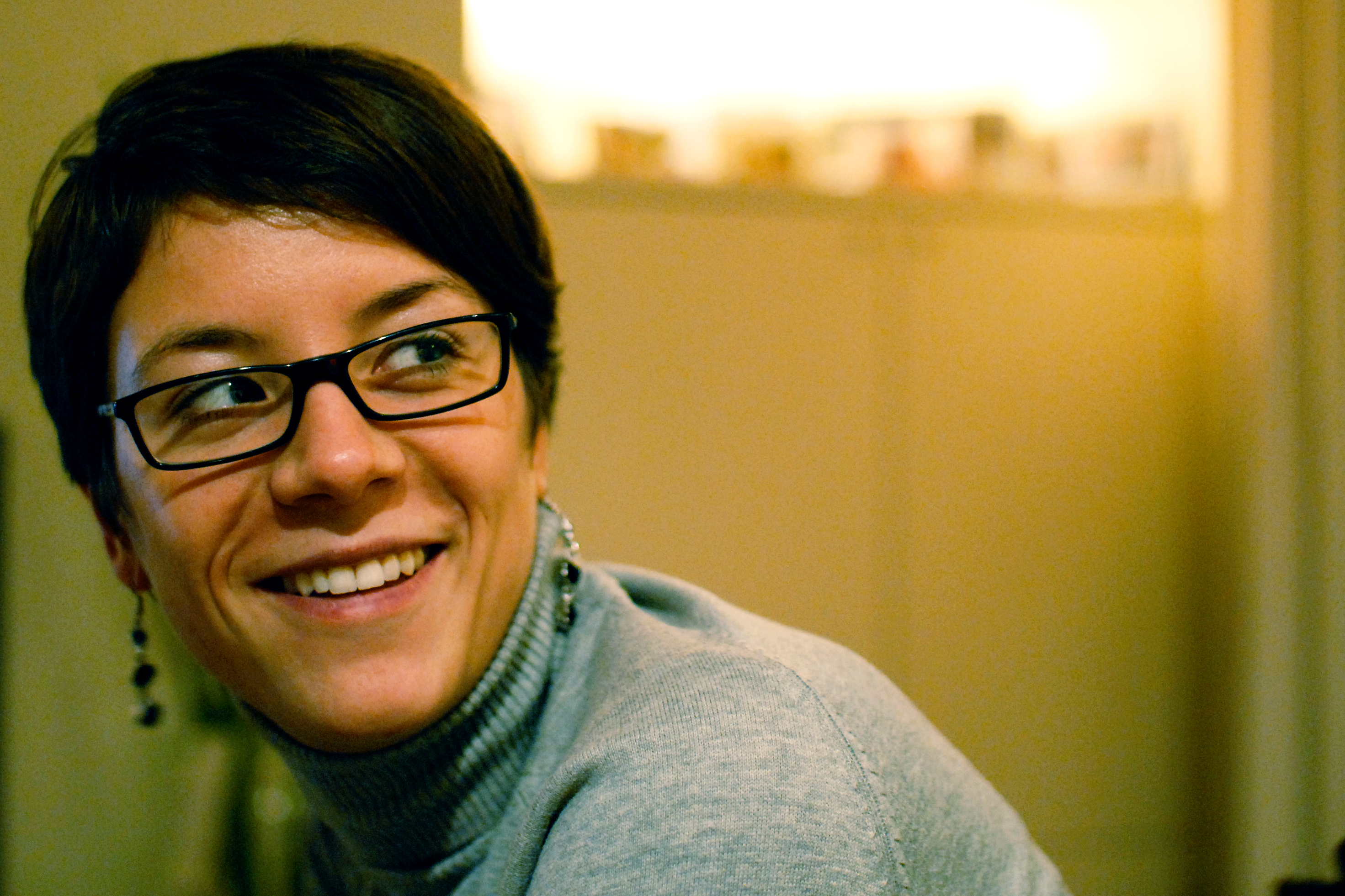 A woman wearing glasses and a gray turtleneck sweater smiles while looking to the side.