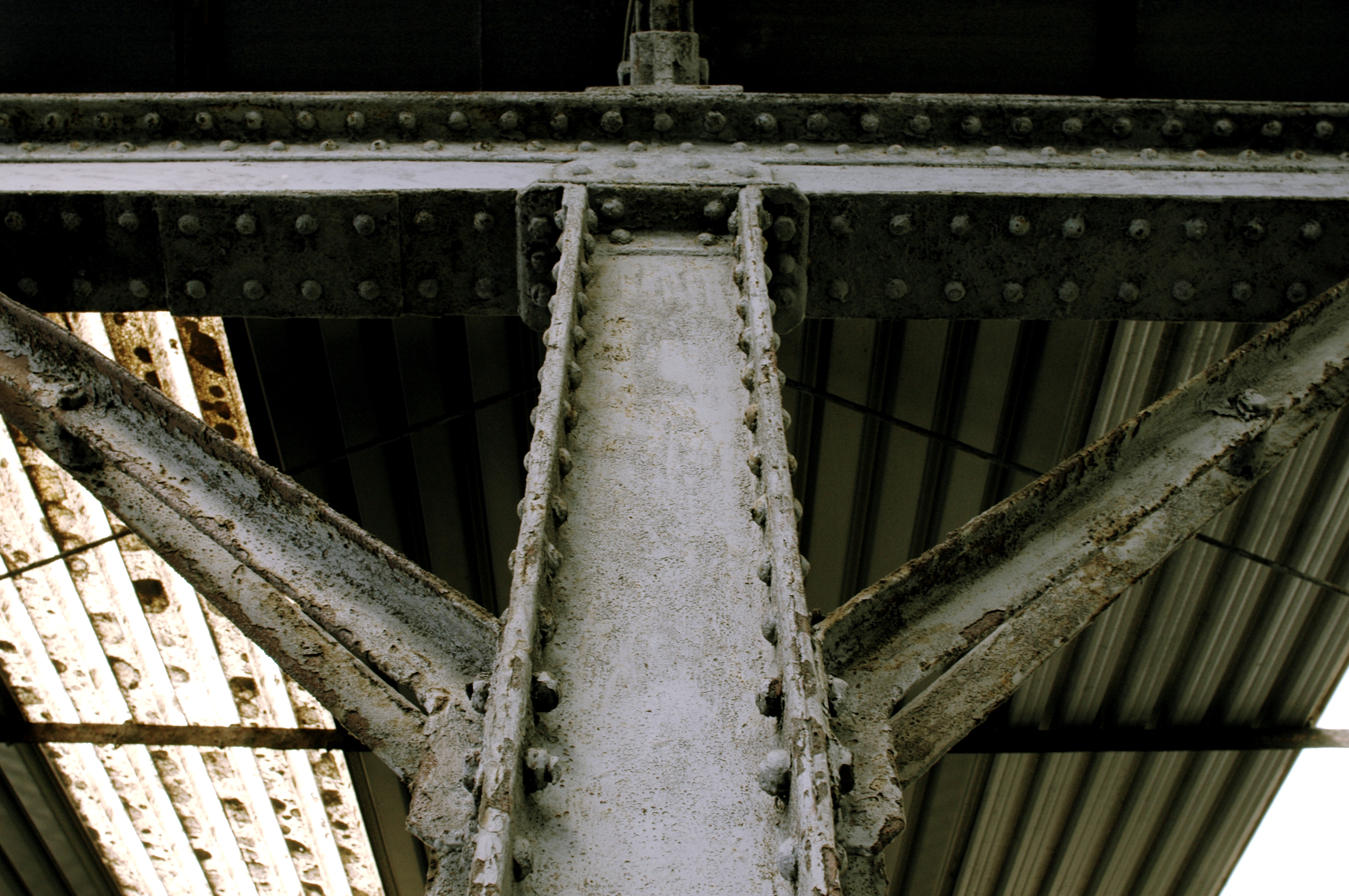 Close-up of a rusted metal beam with rivets, supporting a corrugated metal roof in an industrial structure.