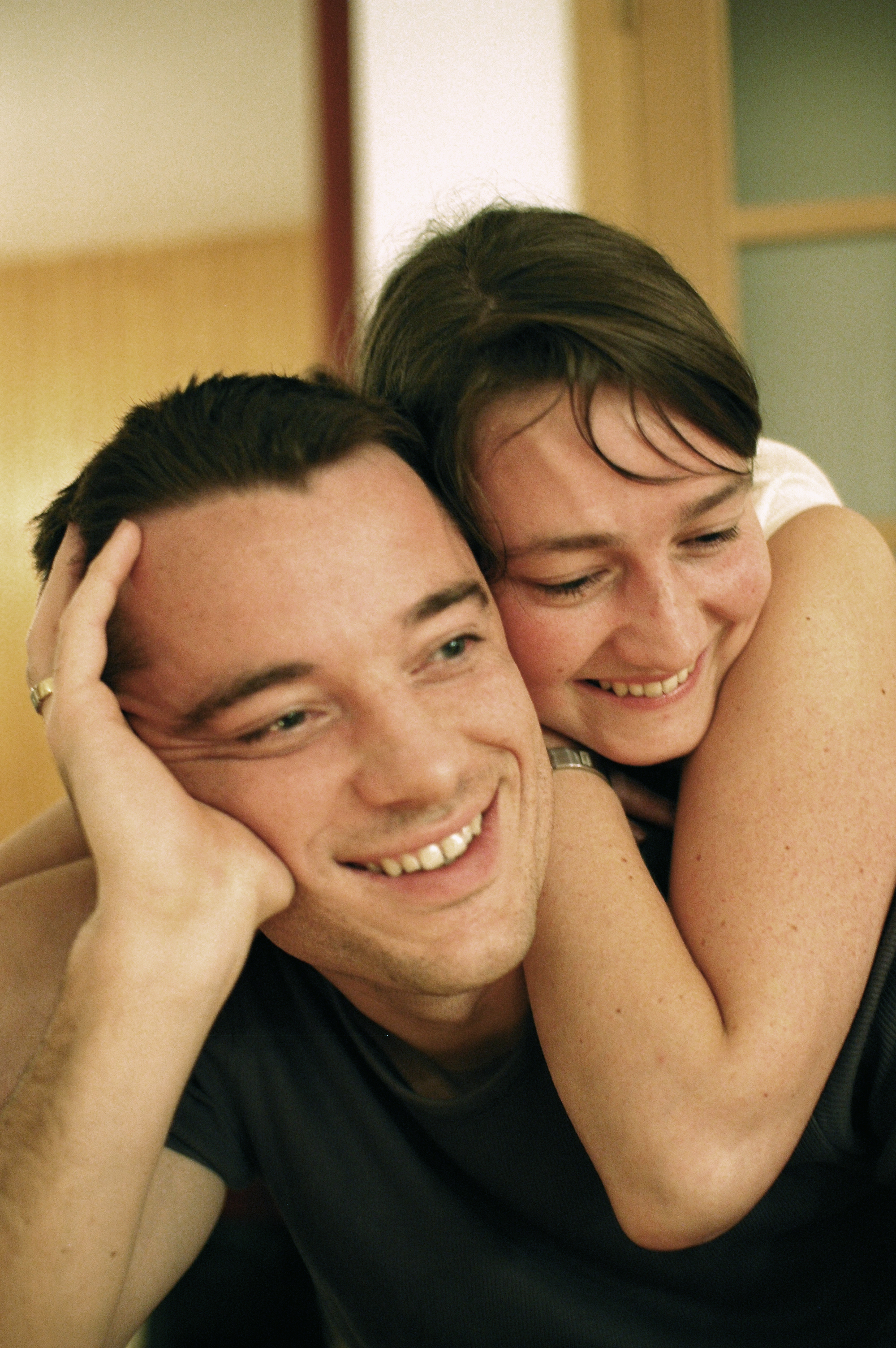 A smiling woman hugs a smiling man from behind, both appearing happy and relaxed indoors.