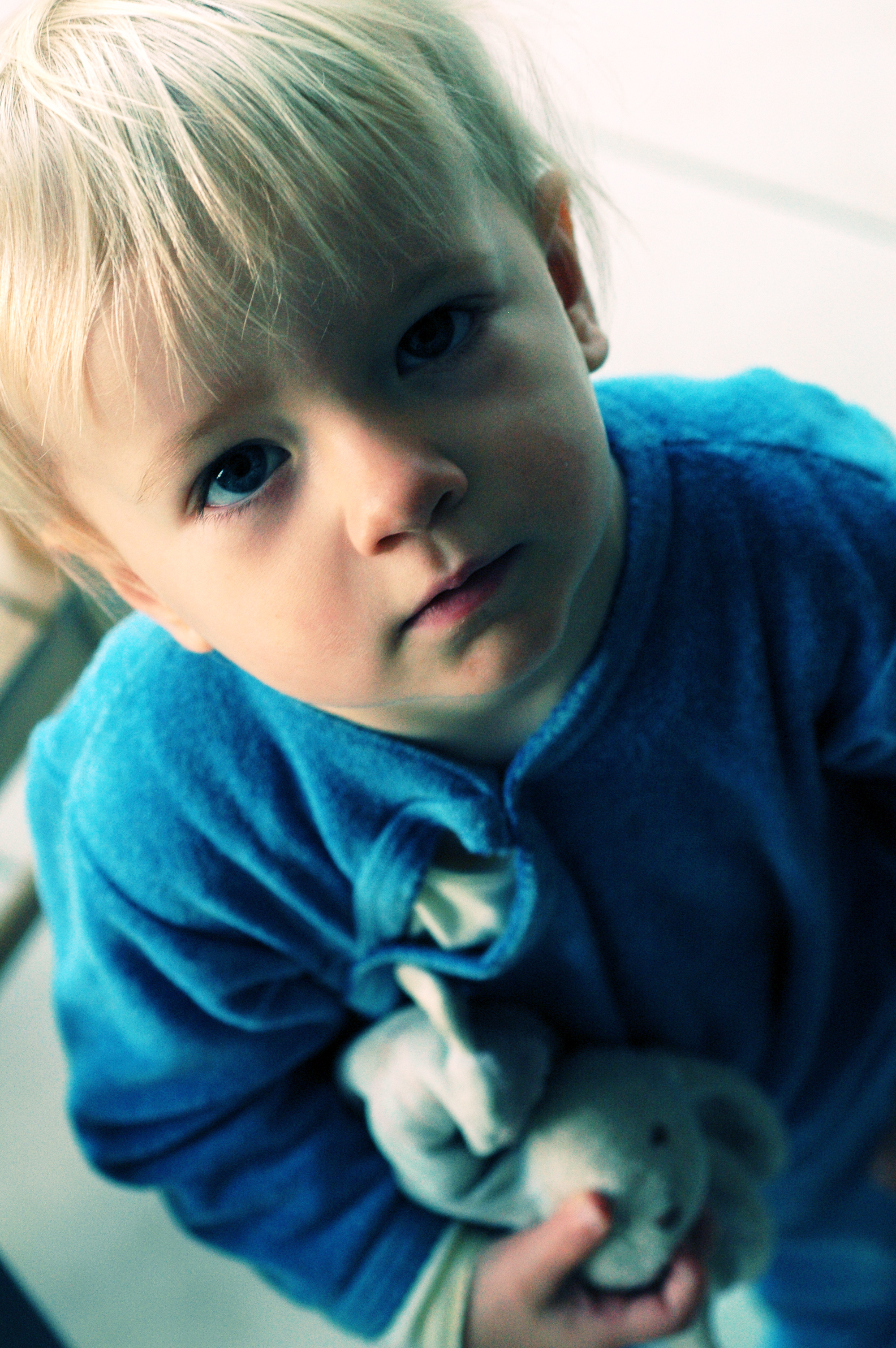 A young child in a blue outfit looks up while holding a stuffed animal close to their chest.