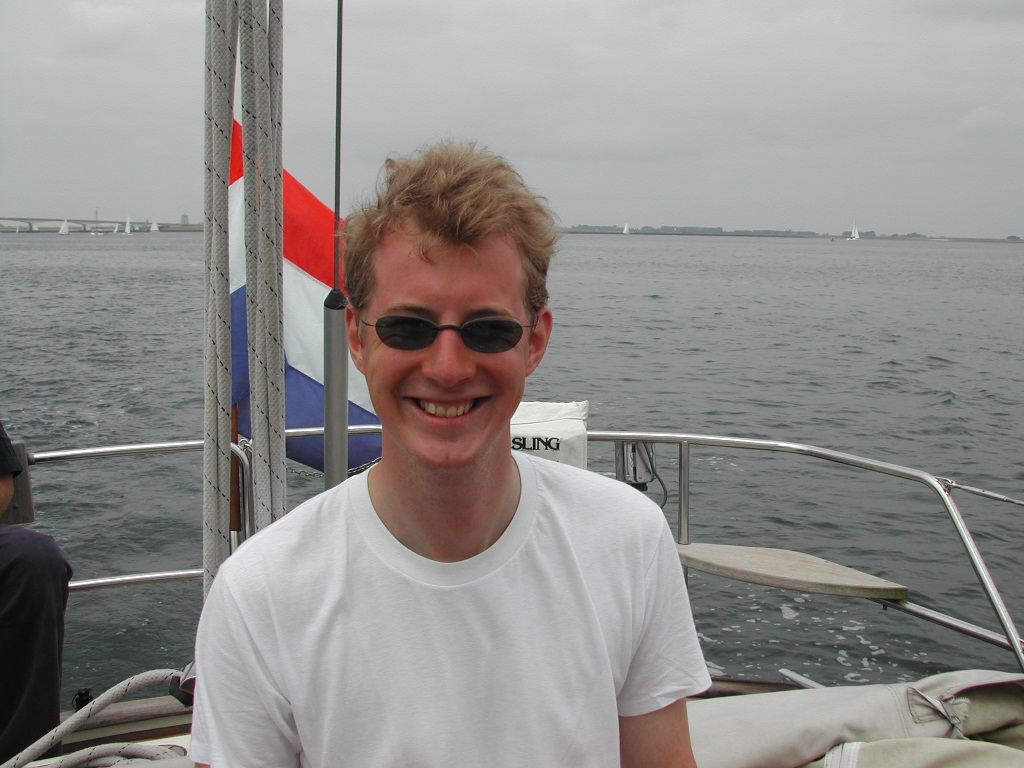 A smiling person wearing sunglasses and a white shirt sits on a boat with water and sailboats in the background.
