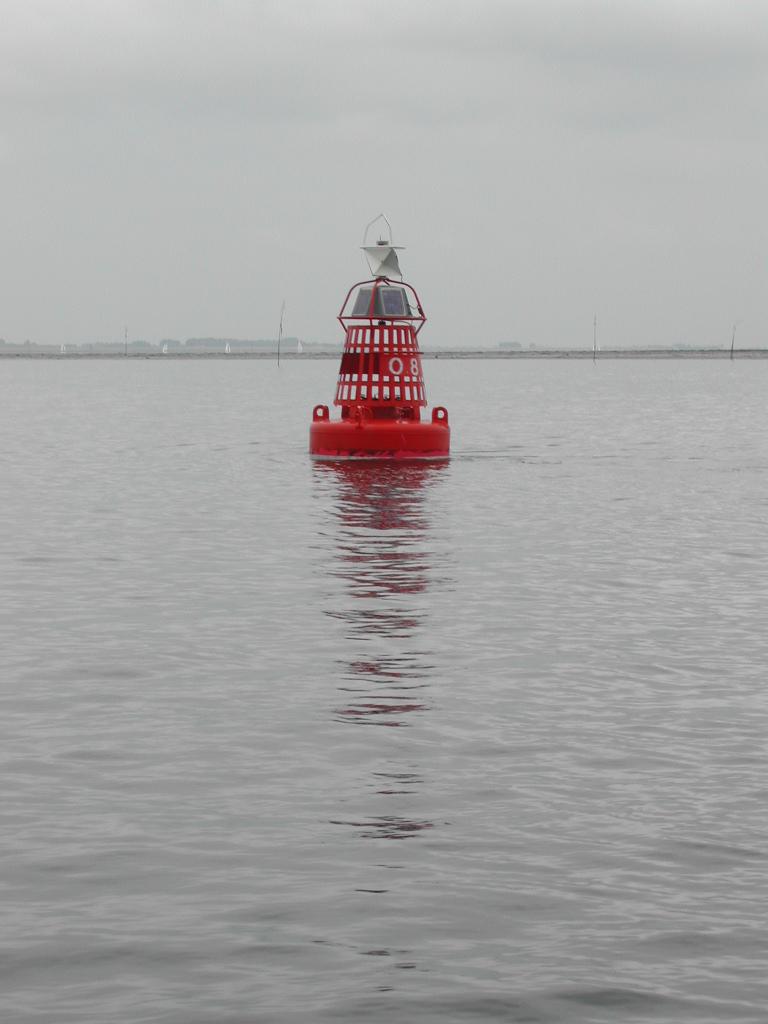 A red navigation buoy floats on calm water, reflecting its shape on the surface under an overcast sky.