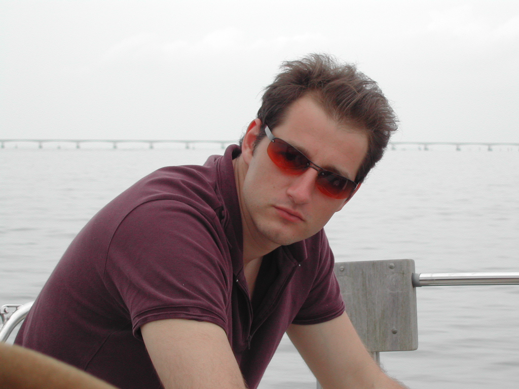 A man wearing red-tinted sunglasses and a maroon shirt sits on a boat with water and a bridge behind him.
