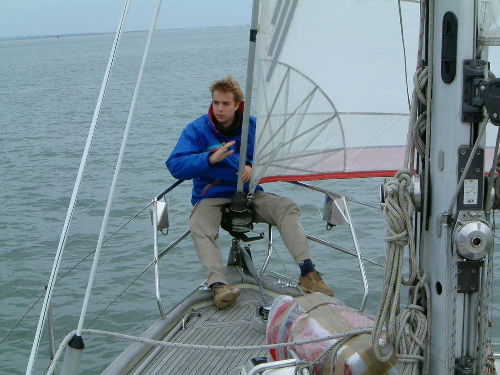 A person in a blue jacket sits on a sailboat, adjusting a rope near the sail.