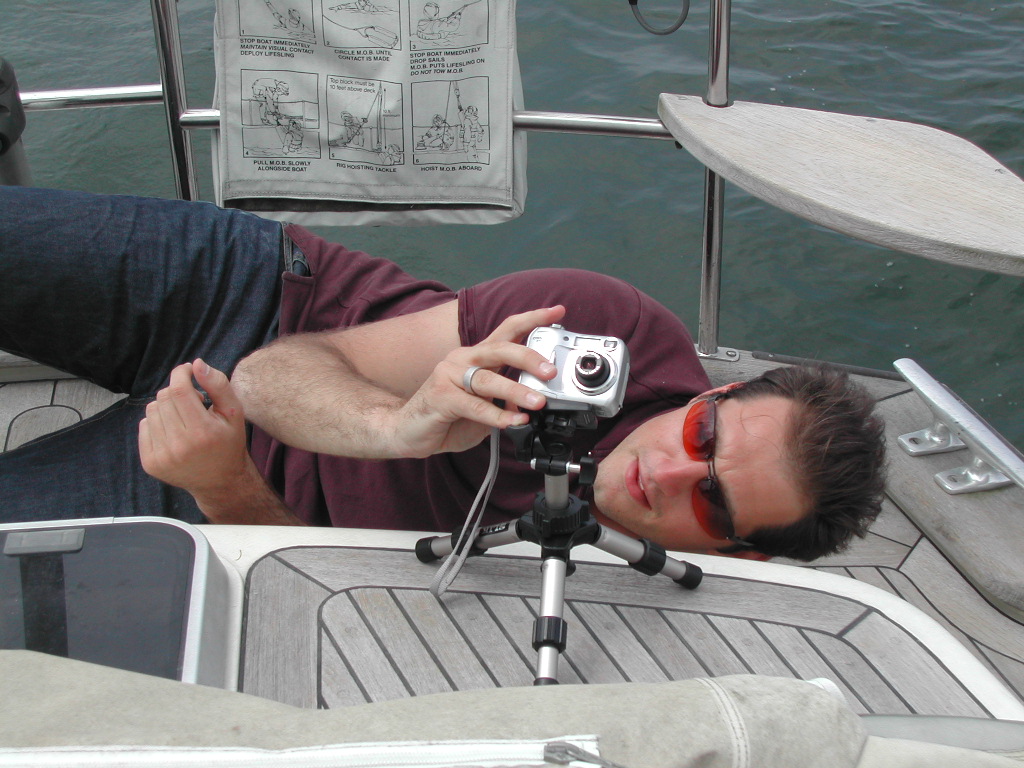 A man wearing sunglasses lies on a boat's deck, adjusting a digital camera mounted on a small tripod.