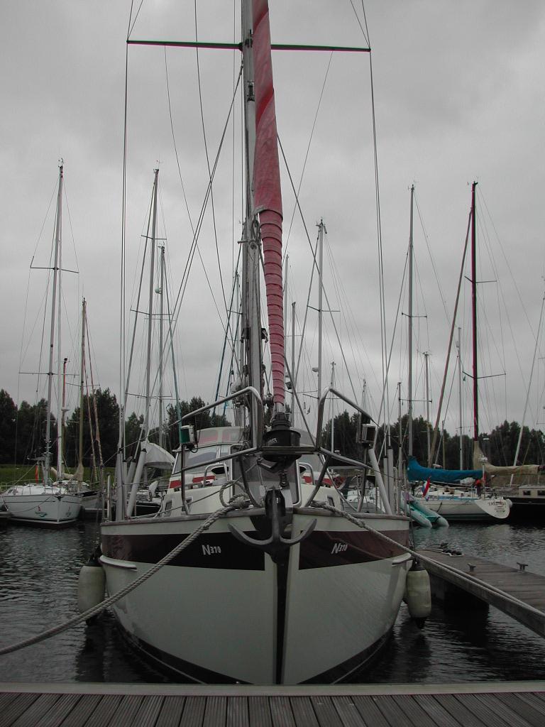 A sailboat with a red mast cover is docked at a marina, surrounded by other boats under a cloudy sky.