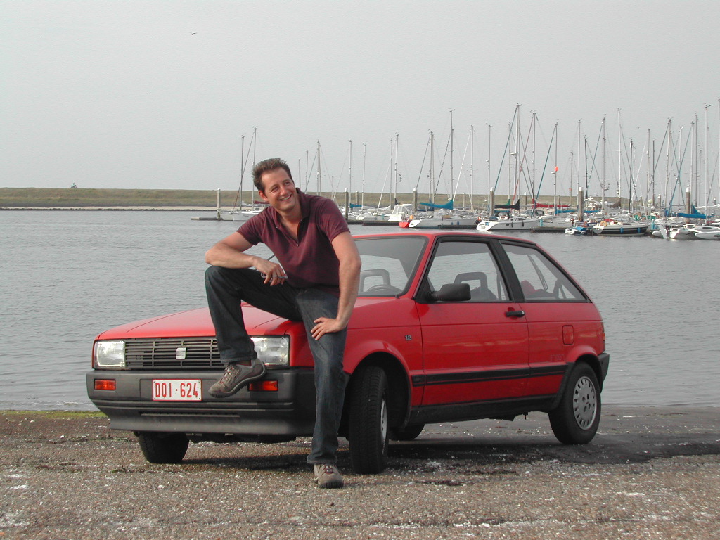 A man sits on the hood of a red car, smiling, with a marina full of boats in the background.