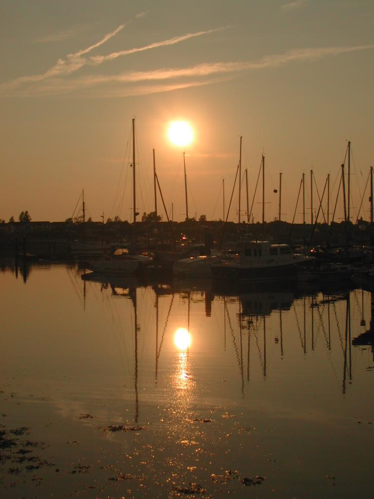 The sun sets over a marina, casting reflections of sailboats and masts on the calm water.