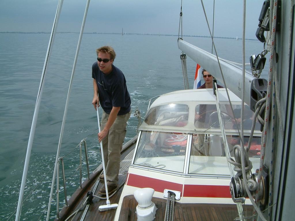 A man wearing sunglasses scrubs the deck of a sailboat while another person steers in calm waters.