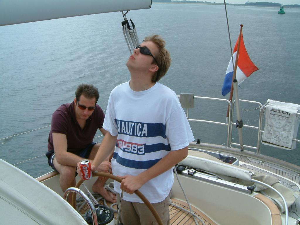 Two men on a sailboat; one steering while looking up, the other sitting and holding a soda can.
