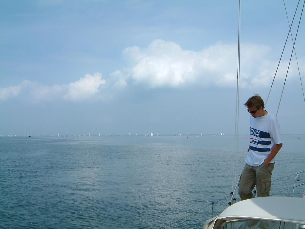 A person wearing sunglasses stands on a boat, looking down, with calm water and distant sailboats in the background.