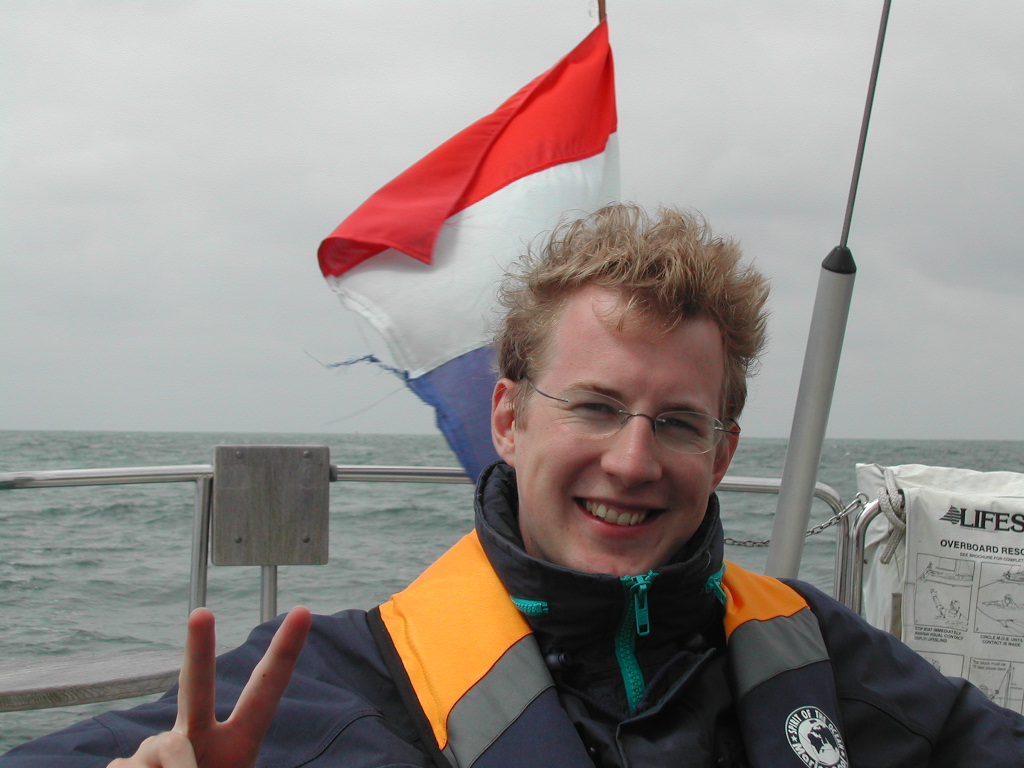 A smiling person wearing a life jacket makes a peace sign while sitting on a boat with a flag behind them.