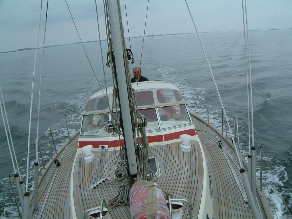 A person steers a sailboat on calm waters, viewed from the deck with rigging and ropes in the foreground.