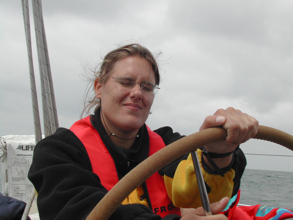 A person wearing a life jacket steers a boat, squinting against the wind under a cloudy sky.