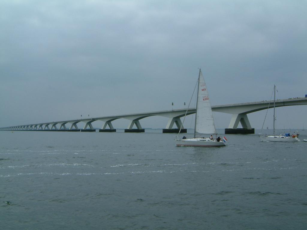 A sailboat moves across the water near a long, modern bridge on a cloudy day.