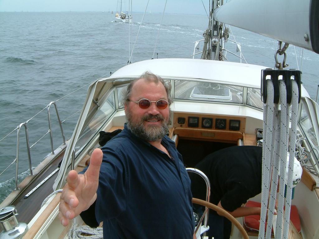 A man with sunglasses and a beard stands on a sailboat, reaching out with his hand and smiling.