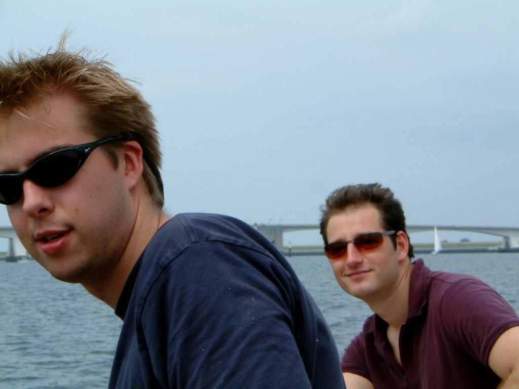 Two men wearing sunglasses sit near the water with a bridge and sailboat in the background.