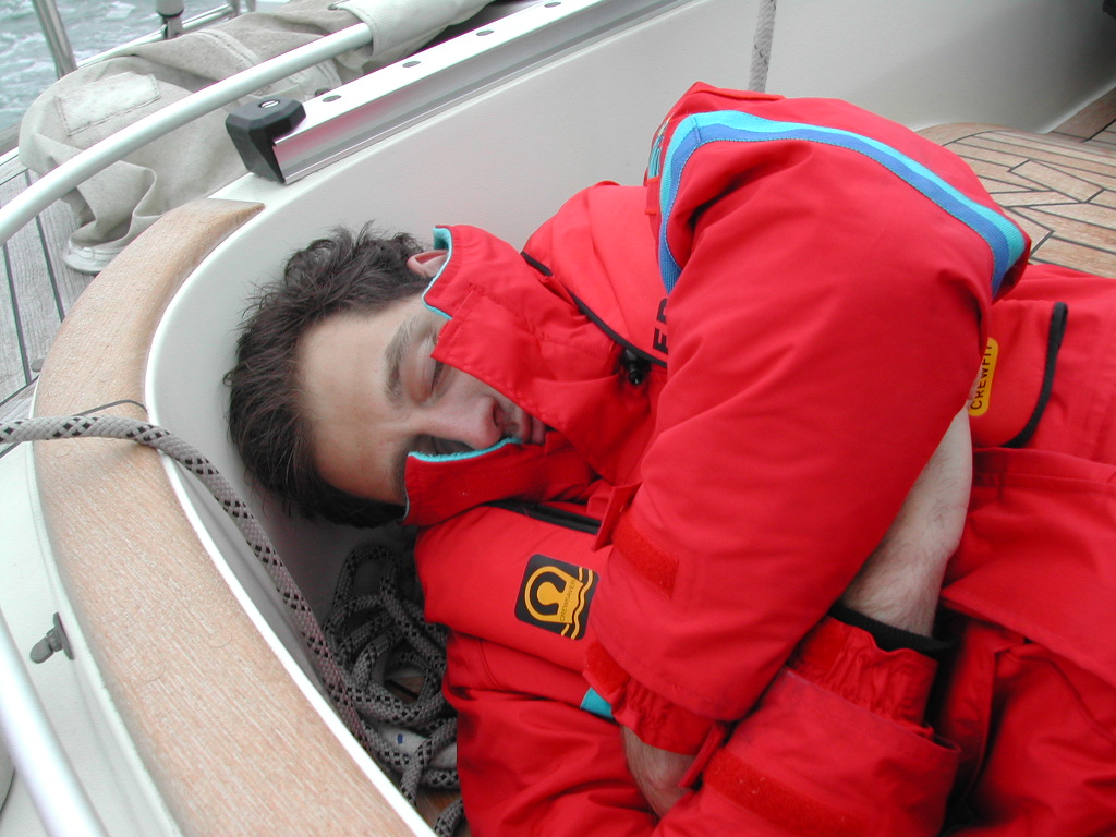 A person in a red waterproof jacket is sleeping curled up on a boat, resting against the side.
