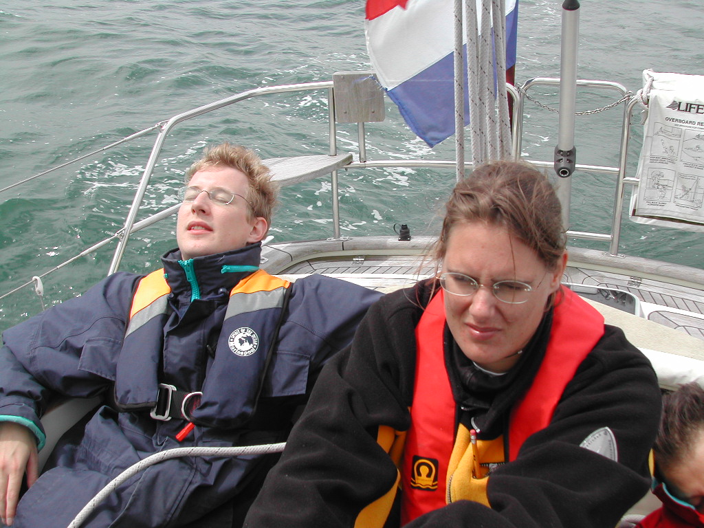 Two people wearing life jackets sit on a boat; one leans back with eyes closed, the other looks forward.