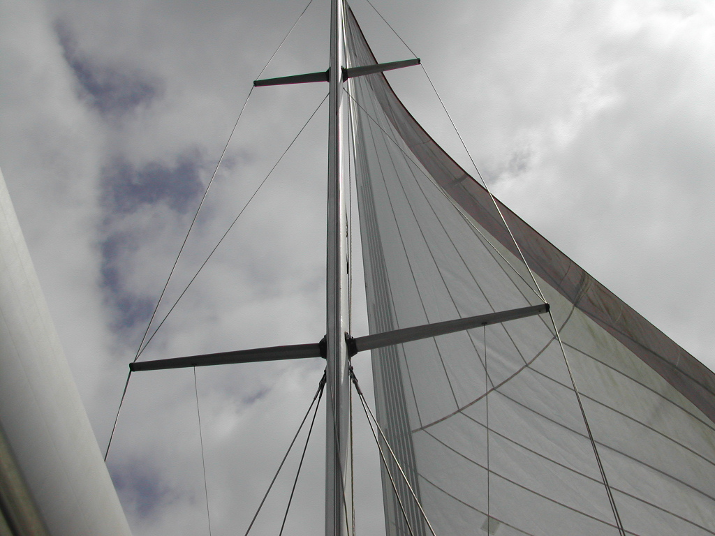 Looking up at a sailboat mast with sails fully extended against a cloudy sky.