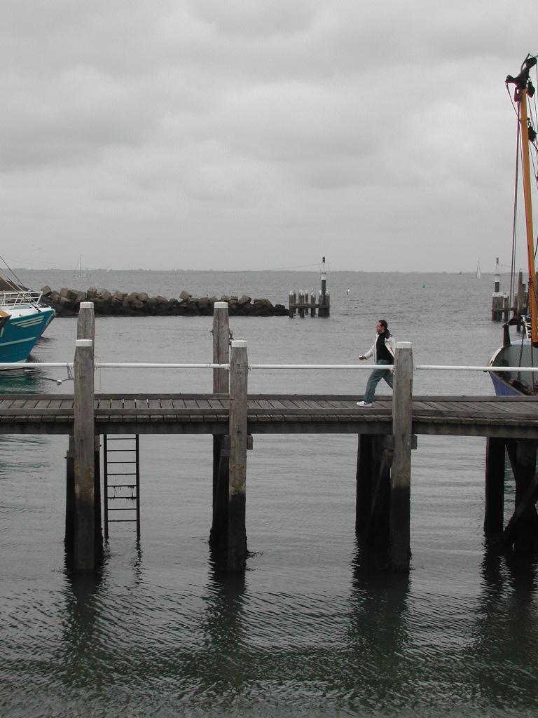 A person walks along a wooden pier with boats nearby and calm water below.