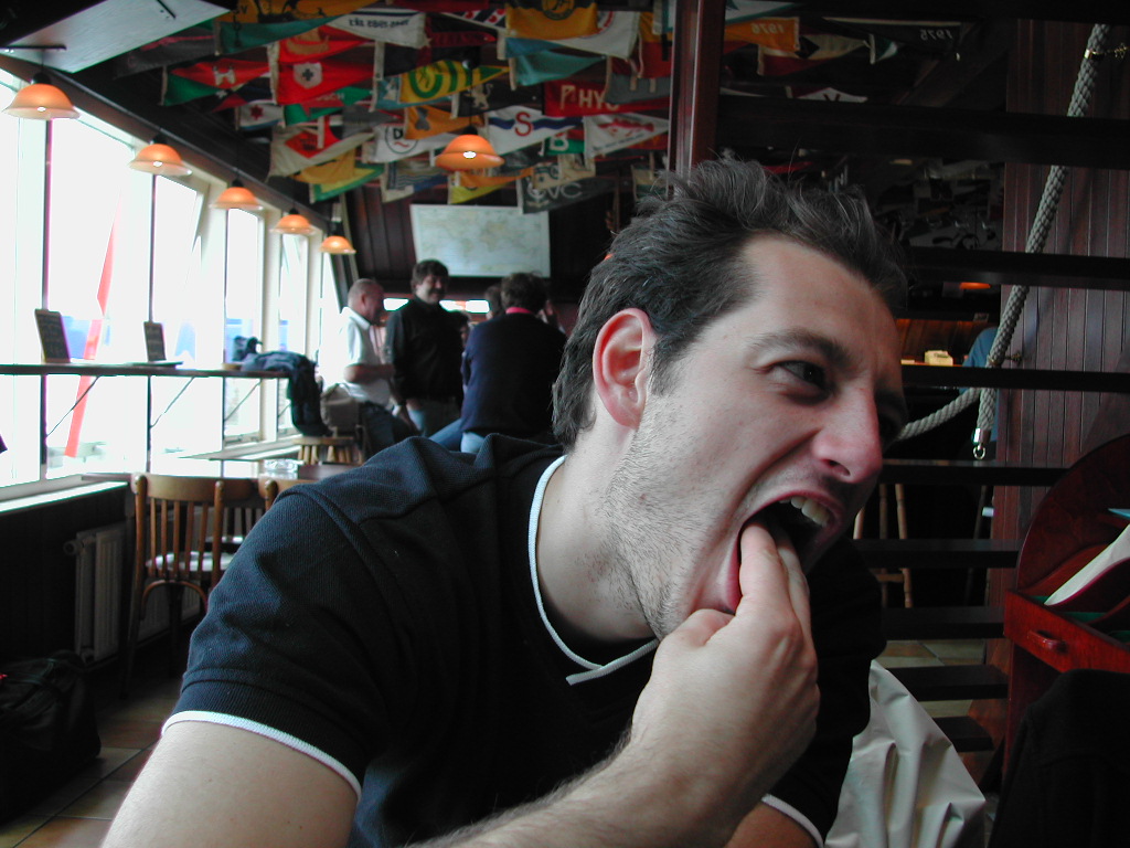 A man in a black shirt sits indoors, sticking his fingers in his mouth with an exaggerated expression.