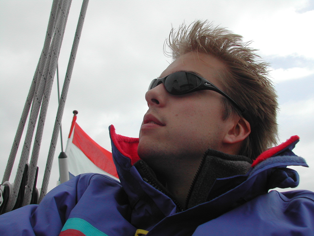 A person wearing sunglasses and a jacket looks up while on a boat, with ropes and a flag behind them.
