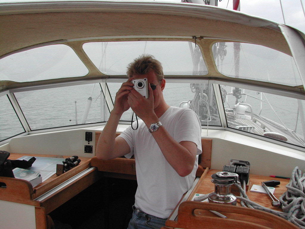 A person in a white shirt and sunglasses takes a photo with a small camera inside a boat's cabin.