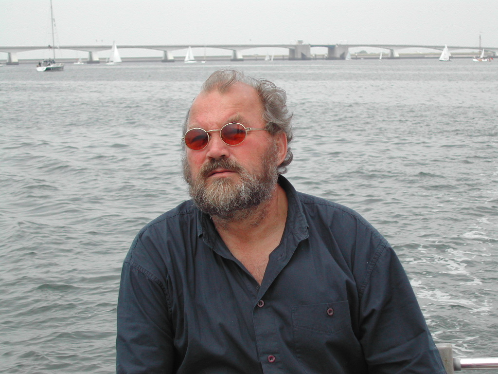 A bearded man wearing red-tinted glasses and a dark shirt sits on a boat with water and a bridge behind him.