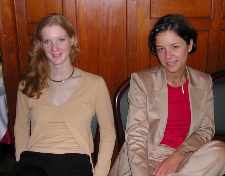 Two women sitting side by side, smiling at the camera, dressed in beige and red outfits in an indoor setting.