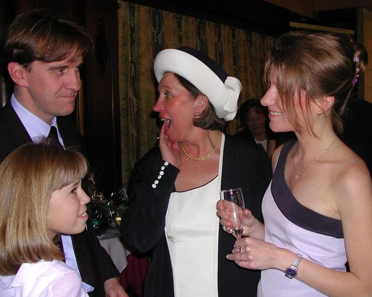 Four people in formal attire are engaged in conversation, with one woman reacting with surprise while holding a drink.