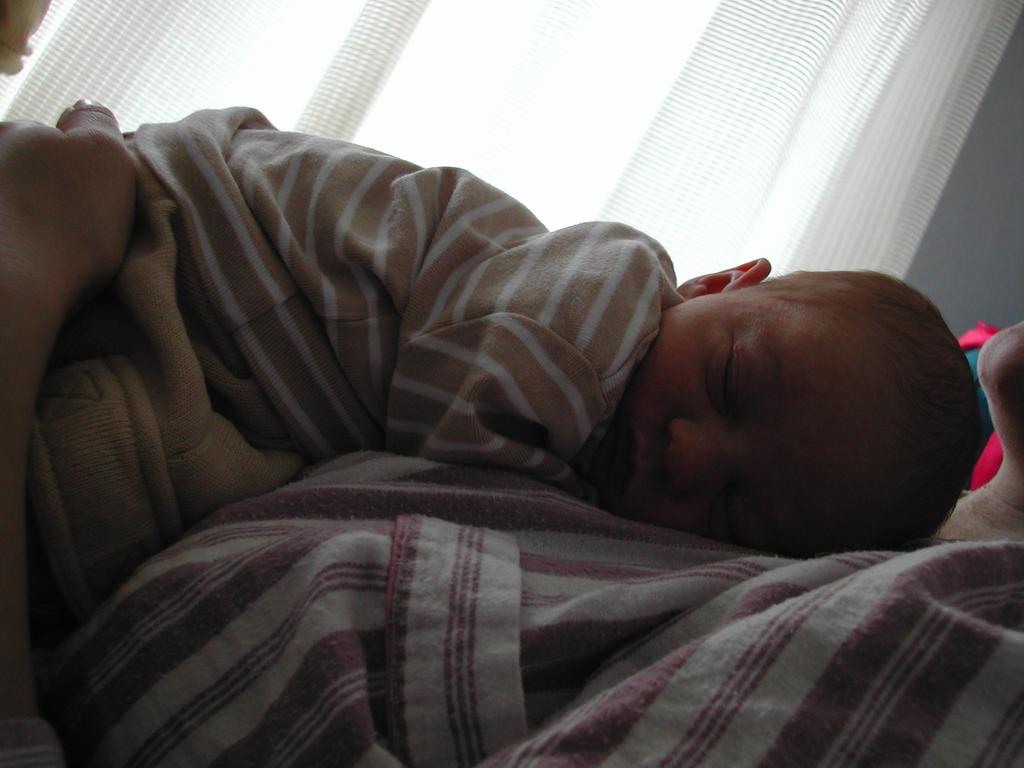 A newborn baby sleeps on a person's chest, dressed in striped clothing, with a hand gently supporting them.