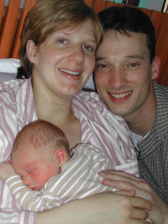A mother holds a sleeping newborn while the father smiles beside them.
