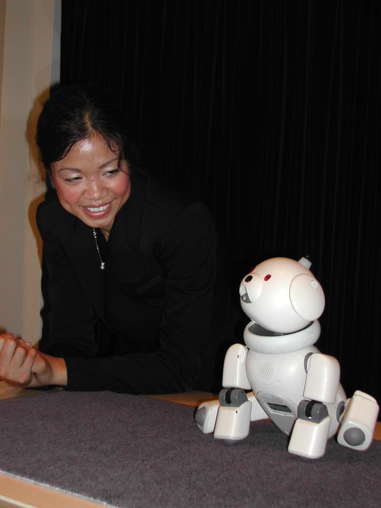 A Sony Aibo robot dog sits on a table while a person nearby watches and smiles.