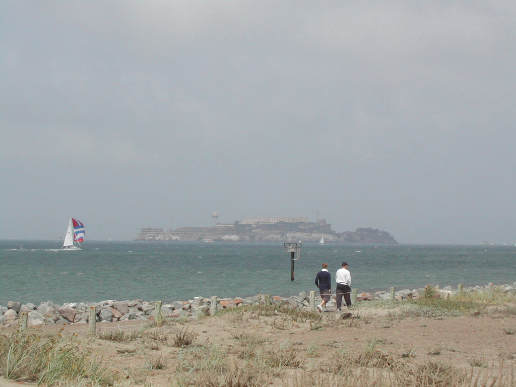 Two people walk along a sandy path with Alcatraz Island and a sailboat visible in the distance.