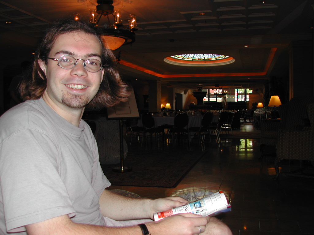 A man with glasses and shoulder-length hair smiles while holding a magazine in a dimly lit restaurant or lounge.