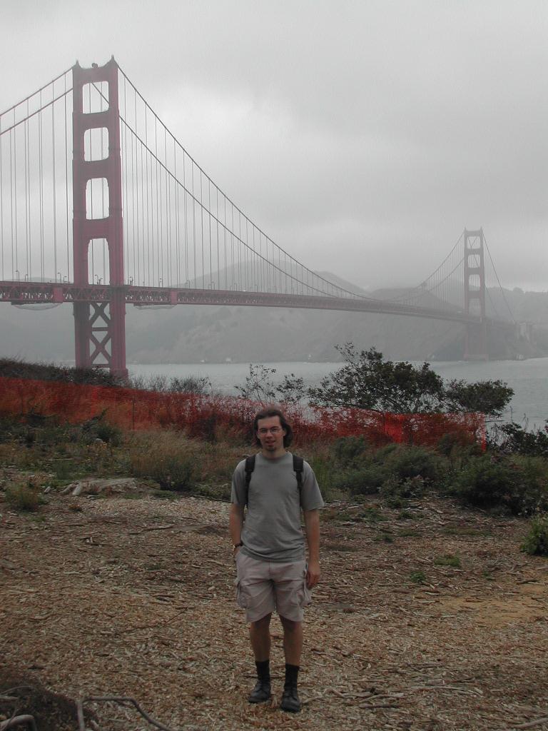 A man wearing a backpack stands in front of the Golden Gate Bridge on a cloudy day.