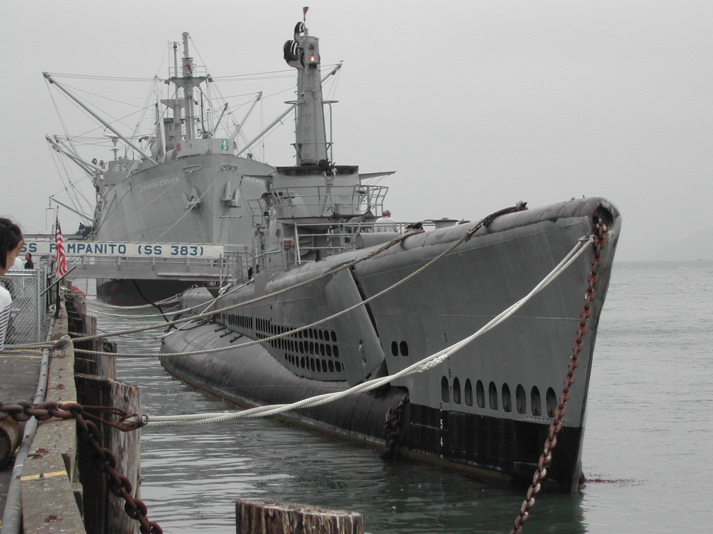 A historic submarine and a large ship are docked at Fisherman's Wharf, with a visitor observing from the pier.