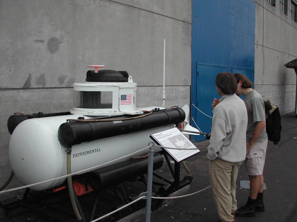 Two people stand near a small experimental submarine, reading an informational sign at Fisherman's Wharf in San Francisco.