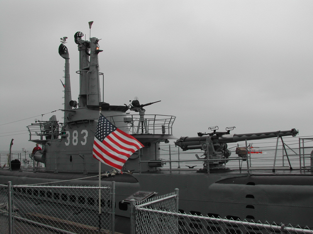 A historic submarine with large deck guns docked behind a fence, with an American flag in the foreground.
