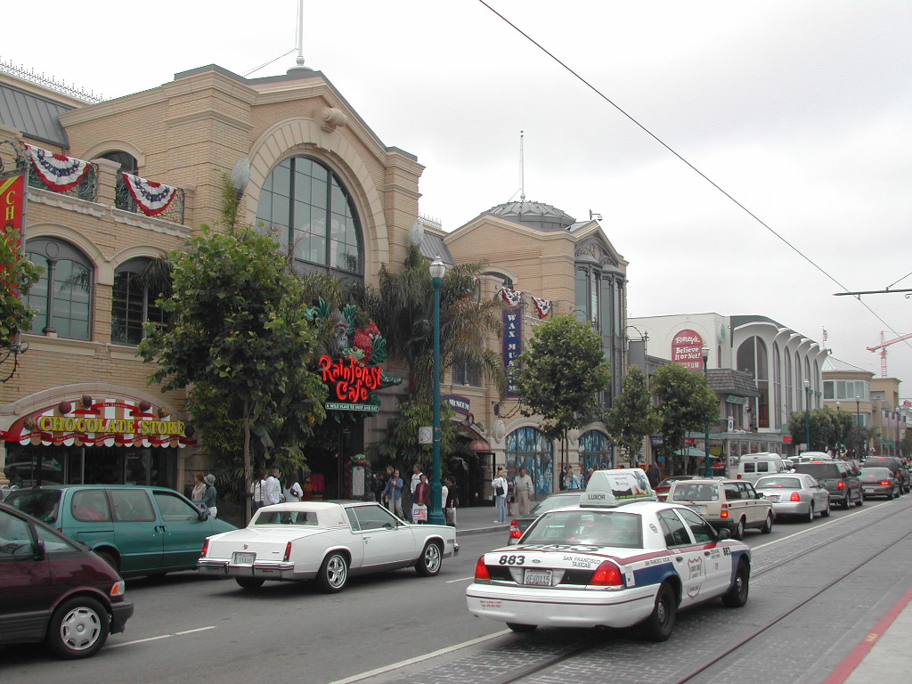 Busy street at Fisherman's Wharf with cars, a taxi, and pedestrians near the Rainforest Cafe and shops.