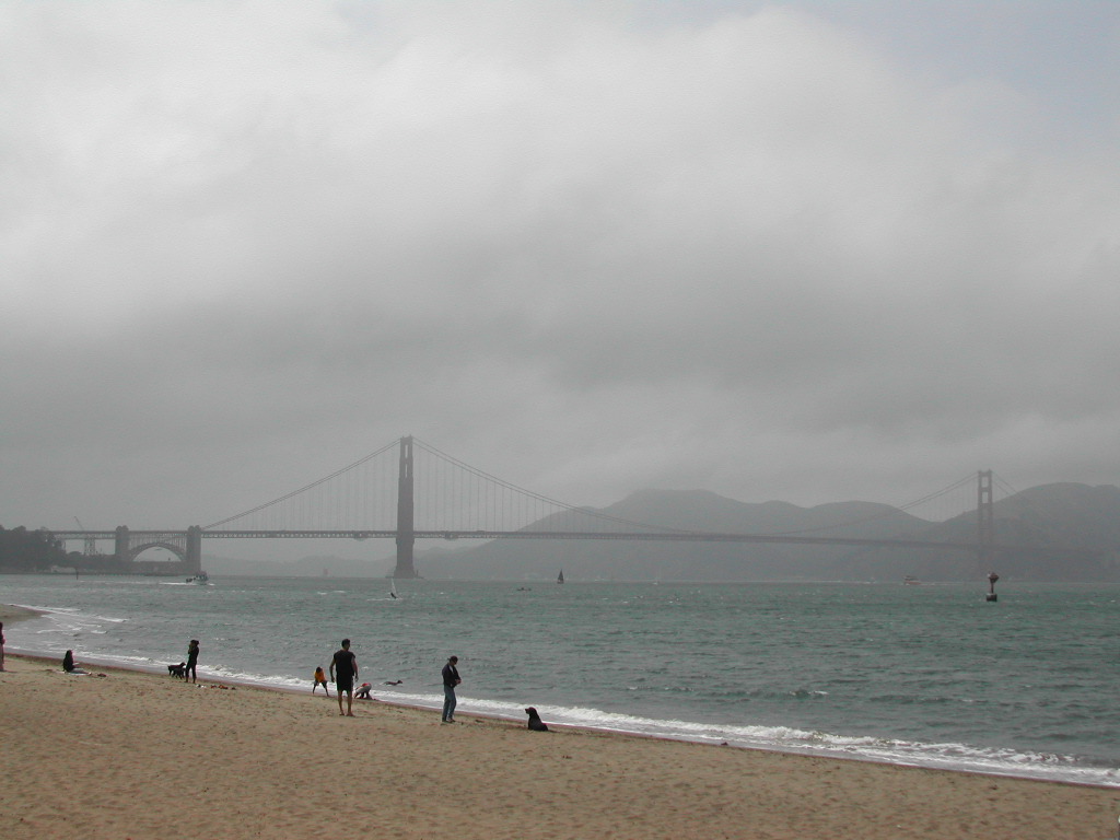People walk along a sandy beach with the Golden Gate Bridge in the background under a cloudy sky.