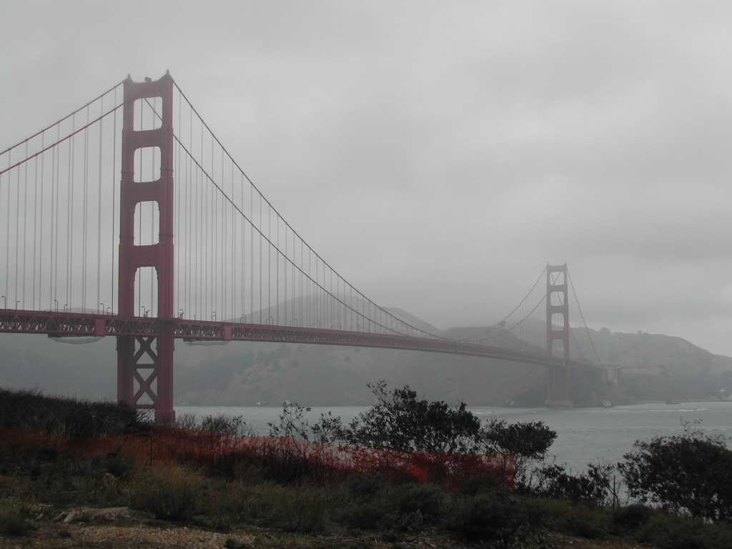 The Golden Gate Bridge stretches across the water on a foggy day, with hills visible in the background.