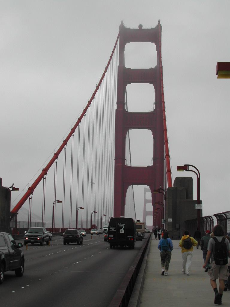 People walk on the sidewalk of the Golden Gate Bridge while cars drive on the road in foggy weather.