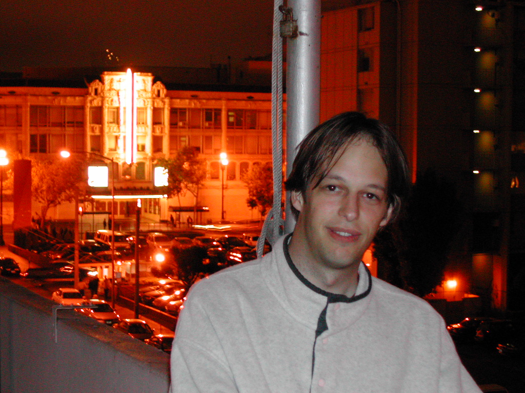 A man in a light-colored jacket sits on a balcony at night with a cityscape in the background.