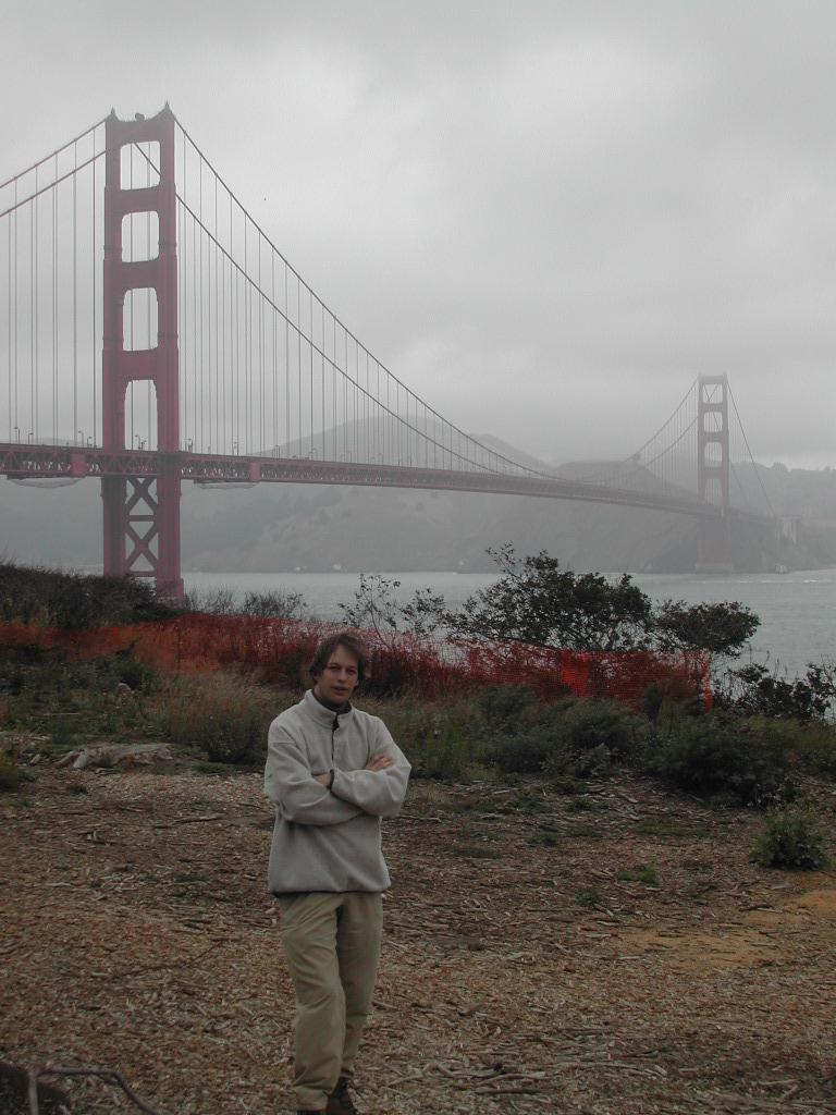 A person in a light jacket stands with arms crossed in front of the Golden Gate Bridge on a cloudy day.
