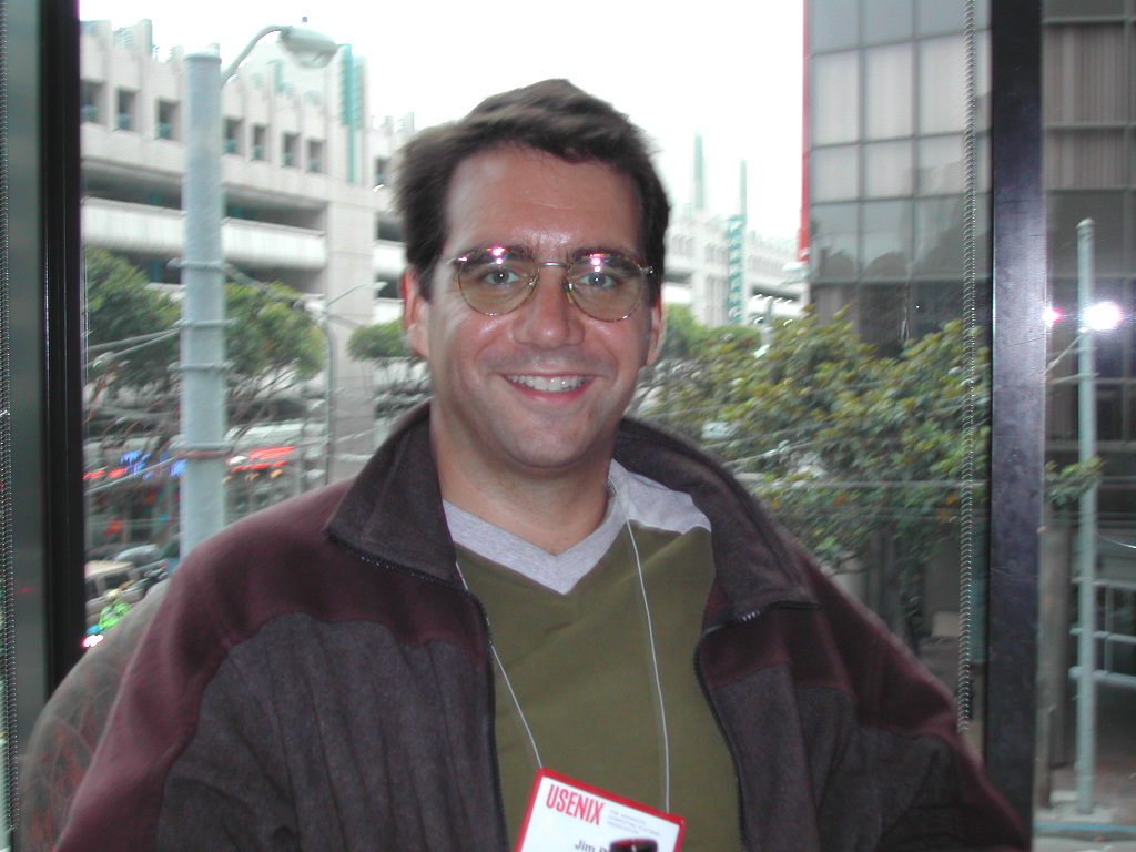 A man wearing glasses and a conference badge smiles while sitting indoors near a window overlooking a city street.